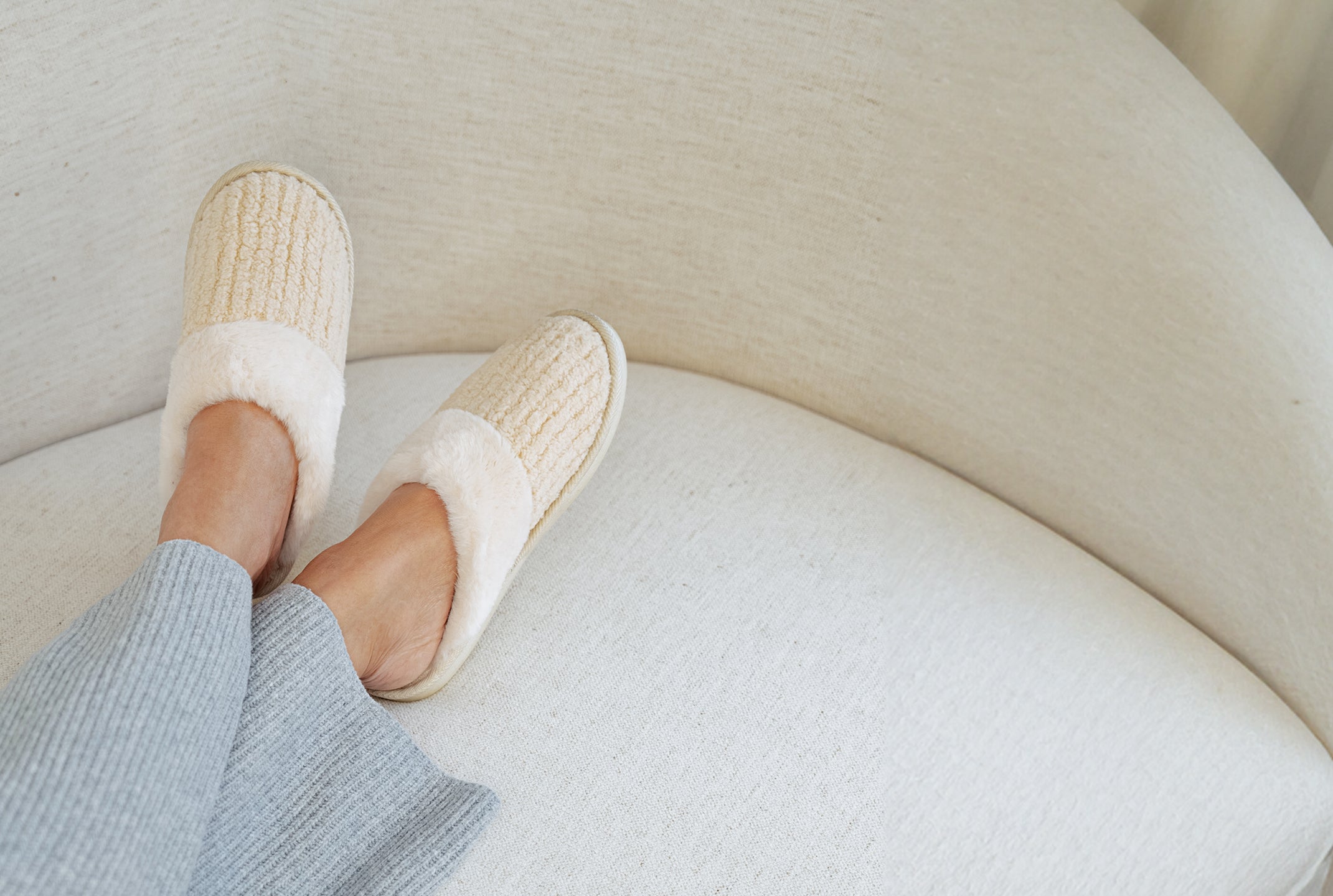 Person wearing white slippers on a light-colored couch. $10 slipper with $99 purchase